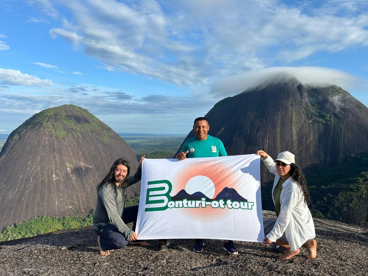 Guía nativo y viajeros con la bandera de Bonturi-ot tour en la cima, frente a los Cerros de Mavicure